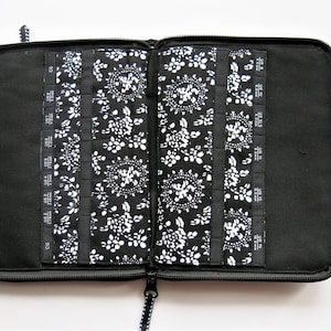 Peut inclure: Étui noir zippé pour aiguilles à tricoter avec un intérieur à motifs floraux. L'étui comporte plusieurs fentes pour les aiguilles et la marque "ChiaoGoo" est visible sur le côté. Le tissu intérieur est un imprimé floral blanc sur fond noir.