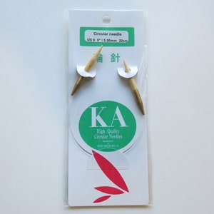 May include: A pack of circular knitting needles, size US 9 (5.50 mm) and 22 cm long. The needles are gold-coloured and have a wooden shaft. The packaging is white with green and red accents. It says "KA High Quality Circular Needles" and "金針" in Japanese.