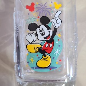 May include: Clear glass tumbler featuring Mickey Mouse in a celebratory pose, with a red shirt, yellow shoes, and white gloves. The design includes colorful confetti and fireworks. The year "2000" is printed at the bottom. © Disney.