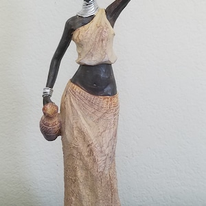 May include: A decorative figurine of a woman carrying a basket on her head and a jug in her hand. The figurine is painted in earth tones, with silver accents on the arms and neck. The woman is wearing a draped dress.