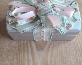 Puede incluir: Una pequeña caja rectangular de madera gris con un lazo decorativo. El lazo está hecho de cintas transparentes de color azul claro y rosa con detalles florales y dorados. La caja está atada con una gran cinta de raso rosa.