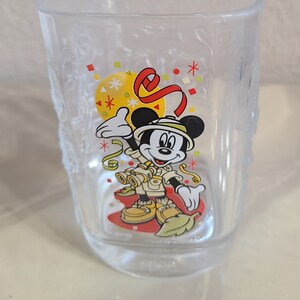 May include: Clear glass tumbler featuring a colorful graphic of Mickey Mouse in safari attire. The design includes a yellow circle, red ribbon, and confetti. The tumbler has a textured exterior and the Disney logo.