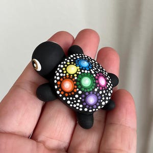 Peut inclure: Une petite figurine de tortue noire avec une finition mate lisse. La carapace présente un motif de points détaillé de style mandala dans des couleurs vives dont jaune, bleu, orange, vert, rose et violet, entouré de nombreux petits points blancs.