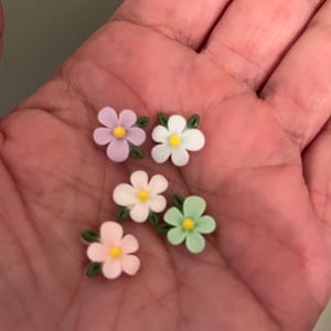 May include: Five small, colorful flower embellishments. Each flower has five petals in pastel shades of pink, white, green, and lavender, with a yellow center. Green leaves are attached to the flowers. These are likely for crafts or decorations.