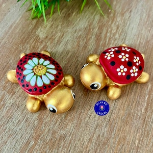 May include: Two small turtle figurines with metallic gold bodies and heads. The turtle on the left features a red shell with a large white daisy design. The turtle on the right has a red shell decorated with white flowers and black dots.