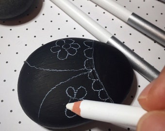 May include: A black stone with a white pencil drawing of a flower design. The pencil is being used to outline the design.