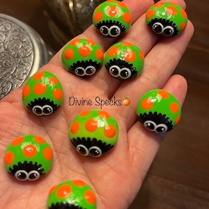 May include: Hand-painted, round, green ladybug-themed stones with orange spots and black detailing. Each stone has large, cartoonish eyes. The text "Divine Specks" is visible in the image.
