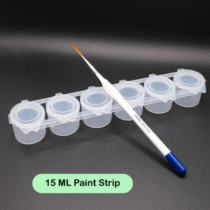 Op de afbeelding: Een doorzichtige plastic verfstrip met zes individuele verfpotjes, elk met een deksel. Een witte penseel met een blauwe handgreep rust op de strip. De tekst "15 ML Paint Strip" wordt weergegeven op een groene rechthoek.