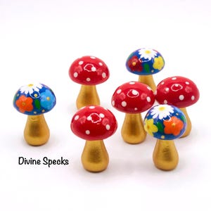 May include: A selection of decorative mushroom ornaments. The mushroom caps are painted red with white spots or blue with flower patterns. The stems are gold-coloured. The text "Divine Specks" is visible.