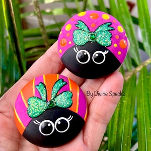 May include: Two painted rocks with ladybug designs. Each rock features a black base, a pink and orange top with a glittery green bow, and white-rimmed eyes. The text "By Divine Specks" is visible.