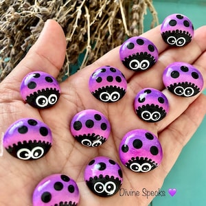 Può includere: Una collezione di dieci pietre dipinte a mano a forma di coccinella. Le pietre sono dipinte in tonalità di viola e rosa con punti neri e occhi. Le pietre sono disposte in una mano.