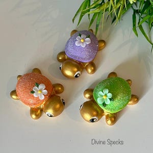 May include: Three decorative turtle figurines with glittery shells in orange, purple, and green. Each turtle has a gold-colored body and a small white flower on its shell. The text "Divine Specks" is visible.