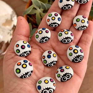 May include: A collection of 11 white ladybug-shaped ornaments with colorful polka dots and black eyes. The ornaments are painted with a variety of colors, including red, yellow, blue, green, and orange.