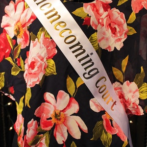 May include: White satin sash with gold lettering that reads "Homecoming Court". The sash is worn over a navy blue dress with a floral pattern.