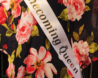 May include: A white sash with gold lettering that reads "Homecoming Queen" on a dark blue floral dress.