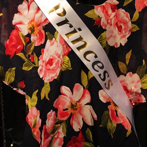 May include: A black floral top with a pink and red flower pattern. A white sash with the word "Princess" in gold lettering is draped across the top. The top is displayed on a mannequin.
