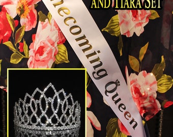 May include: White satin sash with gold lettering that reads "Homecoming Queen" and a silver tiara with rhinestones. The sash and tiara are on a black fabric with a floral pattern.