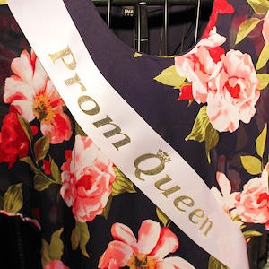 May include: A white satin sash with gold lettering that reads "Prom Queen" on a dark blue dress with a floral pattern.