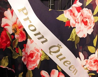 May include: A white satin sash with gold lettering that reads "Prom Queen" on a dark blue dress with a floral pattern.