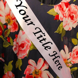 May include: White satin sash with black text that reads "Your Title Here" against a background of a dark blue fabric with a floral pattern.