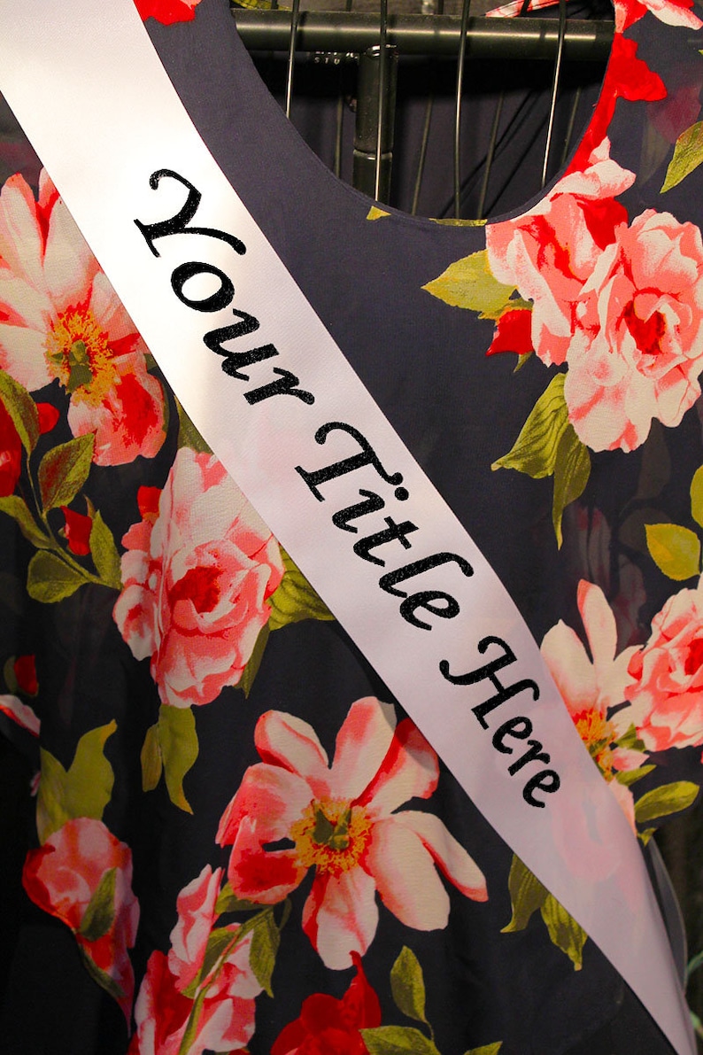 May include: White satin sash with black text that reads "Your Title Here" against a background of a dark blue fabric with a floral pattern.