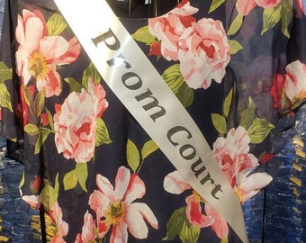 May include: A white satin sash with the text "Prom Court" printed in black. The sash is draped over a navy blue dress with a floral pattern.