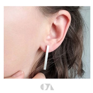 Könnte beinhalten: Ein Paar silberne Ohrstecker in Form von Stäben. Jeder Ohrring ist ein gerader, rechteckiger Stab, der vertikal getragen wird. Die Ohrringe werden an einem Ohr gezeigt. Minimalistisches, modernes Design.