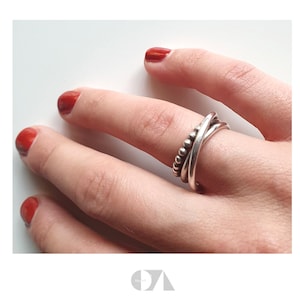 Könnte beinhalten: Ein silberner Ring mit einem geschichteten Design, der ein Band aus kleinen Perlen und zwei sich kreuzende Bänder aufweist. Der Ring wird an einem Finger mit rotem Nagellack getragen.