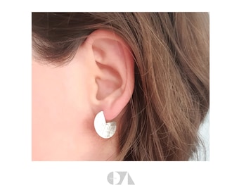 May include: A close-up of a silver earring with a textured, semi-circular design. The earring is worn on an ear, showcasing its unique shape and metallic finish. The earring's surface has a hammered texture, adding visual interest.