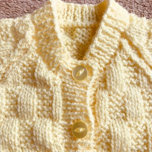 Pu&ograve; includere: Primo piano di un cardigan lavorato a maglia color crema con un motivo strutturato. Il cardigan ha un colletto, due bottoni e un design a trecce. I bottoni sono di colore chiaro e traslucido.