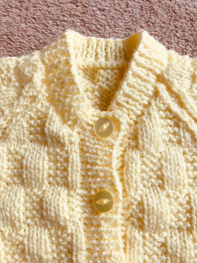 Pu&ograve; includere: Primo piano di un cardigan lavorato a maglia color crema con un motivo strutturato. Il cardigan ha un colletto, due bottoni e un design a trecce. I bottoni sono di colore chiaro e traslucido.