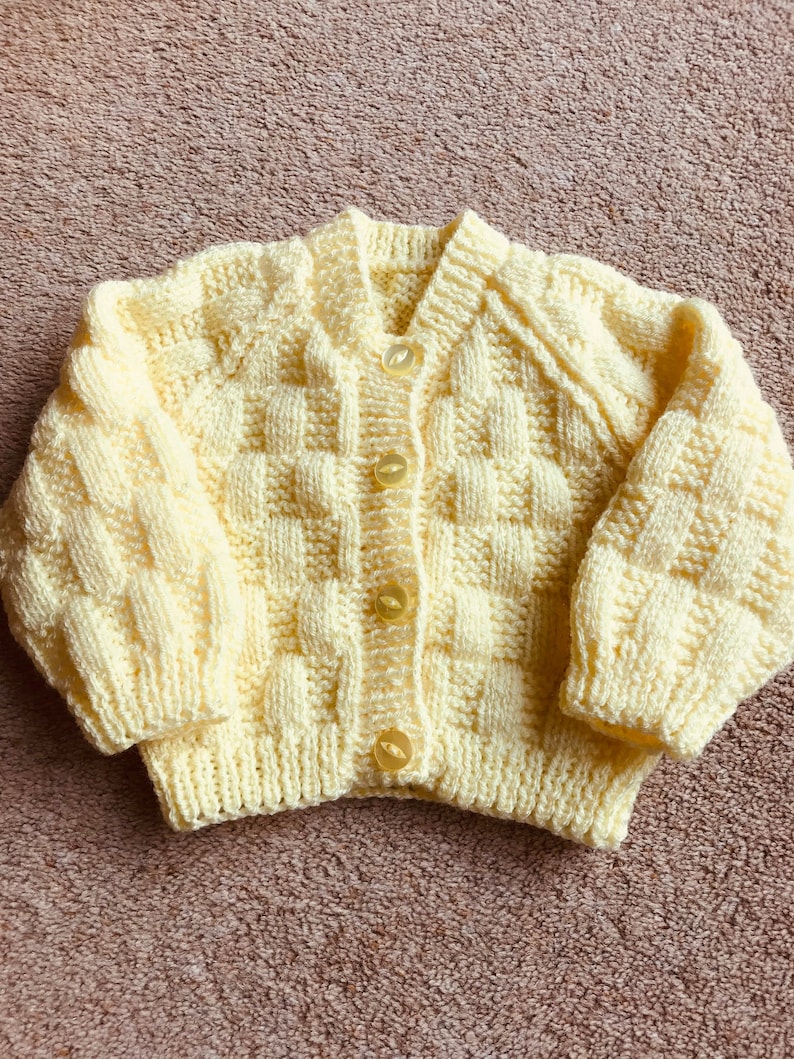 Pu&ograve; includere: Un cardigan per neonati lavorato a mano, giallo chiaro, con un motivo a trama a cestino strutturato. Il maglione ha maniche lunghe, scollo a V ed &egrave; chiuso con tre bottoni gialli. L'indumento &egrave; realizzato in filato morbido.