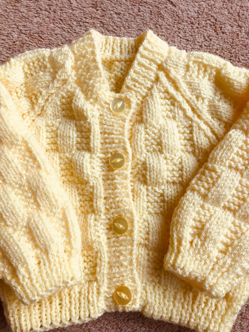 Pu&ograve; includere: Un cardigan giallo chiaro lavorato a mano, con scollo a V e maniche lunghe. Il maglione presenta un motivo a punto strutturato ed &egrave; chiuso con quattro bottoni rotondi e traslucidi. Il colore pastello tenue e il design accogliente suggeriscono un capo per neonati o bambini piccoli.
