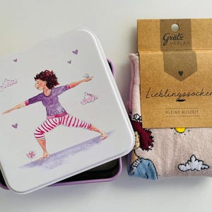 Könnte beinhalten: Eine quadratische weiße Metalldose mit einem Deckel, der eine Aquarellillustration einer Person in Yoga-Pose zeigt, dazu ein Paar hellrosa Socken mit Grafikdruck, verpackt mit einem braunen Pappetikett mit der Aufschrift "Lieblingssocken".