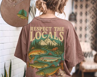 May include: A brown t-shirt with a graphic on the back that says "Respect the Locals" above an illustration of fish in a river with mountains and trees. The front of the shirt has a small graphic of a fish and a tree.
