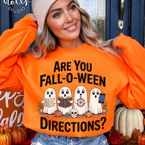 Può includere: Una felpa arancione con la scritta "ARE YOU FALL-O-WEEN DIRECTIONS?" e una grafica di quattro fantasmi, zucche e un teschio. I fantasmi tengono in mano una mappa, un libro, un volante e un telefono. La felpa è indossata da una persona.