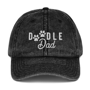 Puede incluir: Gorra de béisbol de mezclilla vintage negra lavada con el texto "Doodle Dad" bordado en blanco y tres huellas de patas.