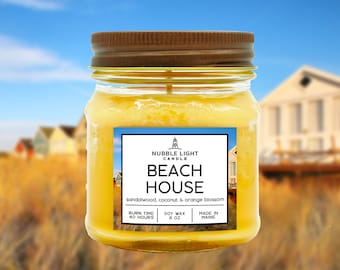 May include: A glass jar candle with a brown lid. The label reads "Nubble Light Candle Beach House sandalwood, coconut, & orange blossom Burn Time 40 Hours Soy Wax 8 oz Made in Maine".