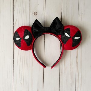 May include: Red and black Deadpool-themed mouse ears headband. The ears feature the character's mask design with white eyes and black accents. A large black bow sits between the ears. The headband is red with black trim.