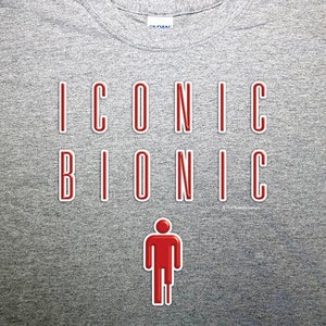 Op de afbeelding: Grijs T-shirt met de tekst "ICONIC BIONIC" in rode letters. Onder de tekst staat een rode stick figure met een beenbrace.