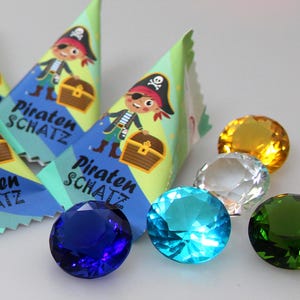Puede incluir: Tres paquetes triangulares de papel con un pirata de dibujos animados sosteniendo un cofre del tesoro y el texto Piraten SCHATZ, acompañados de cuatro gemas de plástico facetadas en colores azul, azul claro, transparente y verde.