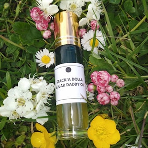Op de afbeelding: Een heldere glazen roll-on fles met een gouden dop en een wit etiket met de tekst "Stack'a Dolla Sugar Daddy Oil". De fles is omgeven door witte, roze en gele wilde bloemen en groen gras.