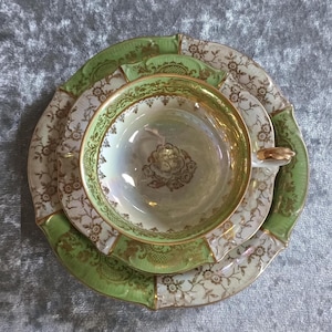 May include: A vintage tea set featuring a cup, saucer, and plate. The cup and plate have a gold rim and a floral design. The saucer and plate have a green border. The cup has a gold handle. The set is on a gray surface.