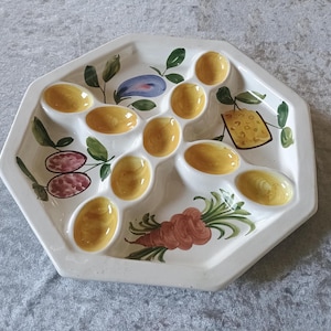 May include: White ceramic serving dish with an octagonal shape. The dish features painted illustrations of food items, including olives, cheese, carrots, and what appear to be deviled eggs. The dish has multiple oval-shaped indentations.