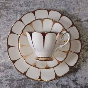 Könnte beinhalten: Ein weißes Tee-Set mit goldenen Verzierungen. Das Set besteht aus einer Tasse, einer Untertasse und einem Teller. Die Tasse und der Teller haben einen gewellten Rand. Das Set steht auf einer grauen Oberfläche.