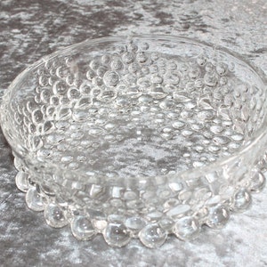 Könnte beinhalten: Eine klare Glasschüssel mit einer strukturierten Oberfläche. Die Schüssel hat eine runde Form und einen erhöhten Rand. Die Schüssel ist mit kleinen, runden Blasen verziert.
