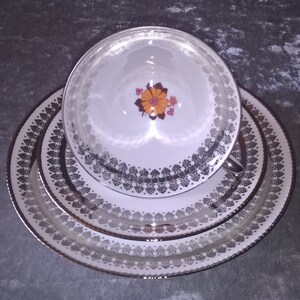 Könnte beinhalten: Ein Set aus drei weißen Keramiktellern mit silbernem Rand und einem Blumendesign. Die Teller sind ineinander verschachtelt, wobei der kleinste Teller oben liegt. Das Blumendesign zeigt eine gelbe Blume mit orangefarbenen und braunen Akzenten.