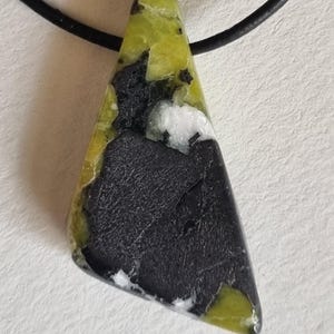Peut inclure: Un collier avec un pendentif triangulaire en pierre. Le pendentif présente une base noire avec des accents blancs et vert-jaune. Le pendentif est attaché à un cordon noir. Bijou unique.