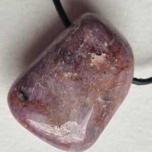 Può includere: Un ciondolo in pietra preziosa lucidata di colore viola. La pietra ha una forma liscia e arrotondata con variazioni naturali di colore, tra cui striature rosso-brunastre e viola più chiare. È infilato su un cordino nero, adatto per essere indossato come collana.