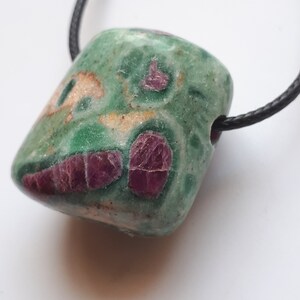 Könnte beinhalten: Ein grüner und rosafarbener Edelstein-Anhänger mit einer schwarzen Schnur. Der Edelstein hat eine raue, natürliche Textur und ist mit rosa und weißen Einschlüssen übersät.